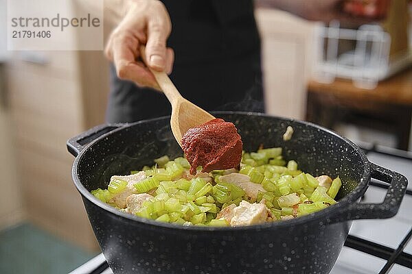 Unrecognizable woman adds tomato paste to a pot filled with diced chicken and chopped celery. to enhance flavour