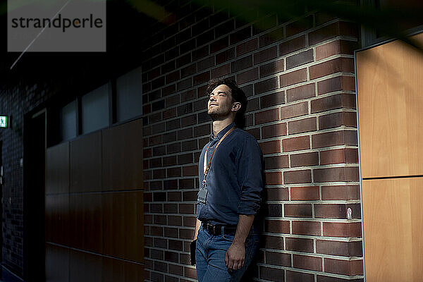 Young businessman with eyes closed standing in front of brick wall