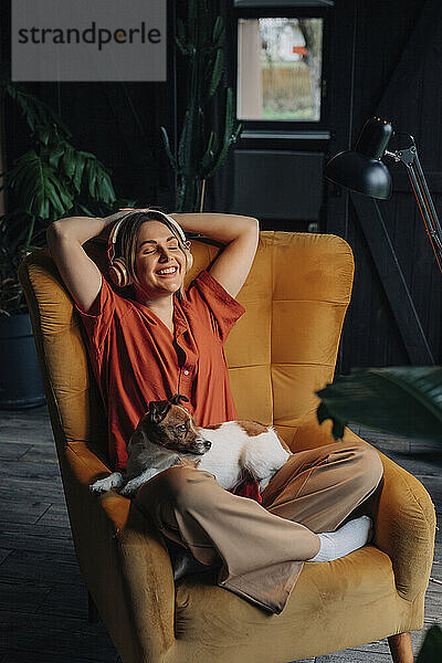 Relaxed young woman wearing wireless headphones listening to music and sitting with dog at home