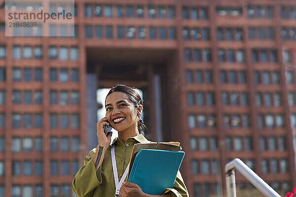 Cheerful businesswoman talking on smart phone near office building