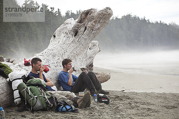 Hikers sit against a giant driftwood log sipping hot tea on a beach along the West Coast Trail  British Columbia  Canada.