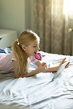 Blondes Mädchen liegt im Bett und leckt Lolli, während sie ein digitales Tablett benutzt