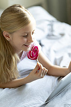 Lächelndes blondes Mädchen liegt mit einem Lolli auf dem Bett und benutzt ein digitales Tablett
